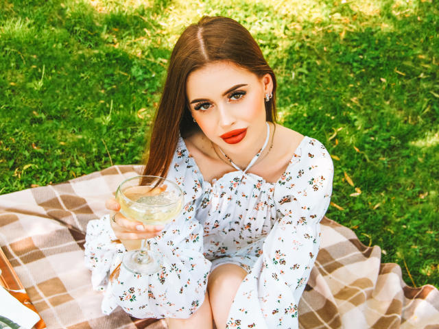 CherylFoxy with long brown hair, red lipstick, floral dress, holding a glass of white wine, sitting on a brown and white checkered blanket in a grassy park.