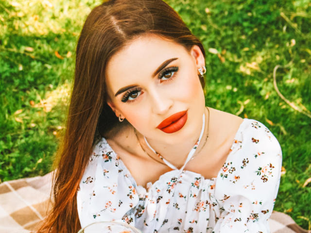 CherylFoxy with long brown hair, red lipstick, and floral dress, looking up with green eyes, small earrings, and a necklace, sitting on a checkered blanket.