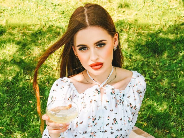 CherylFoxy with long brown hair, wearing a white floral dress, holding a glass of water, has red lipstick, and earrings, sitting on grass, looking at camera.