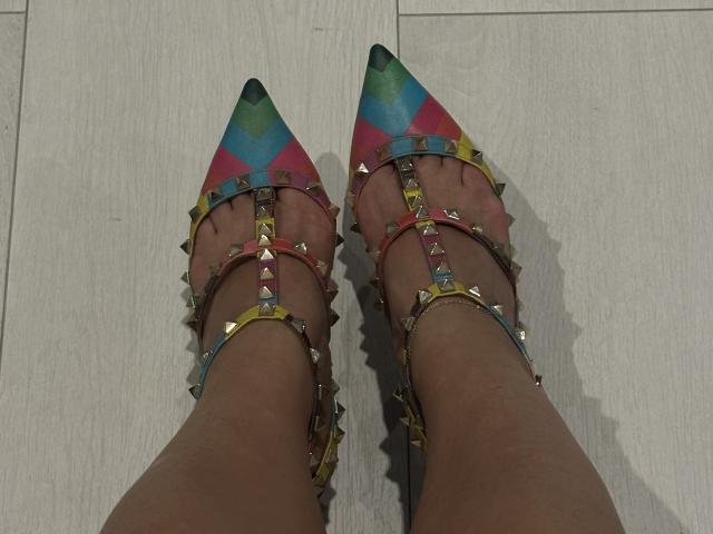 Stefanni's feet wearing colorful, spiked, pointy-toe sandals with rainbow patterns. Her legs are tan, and the sandals have metal studs and straps.