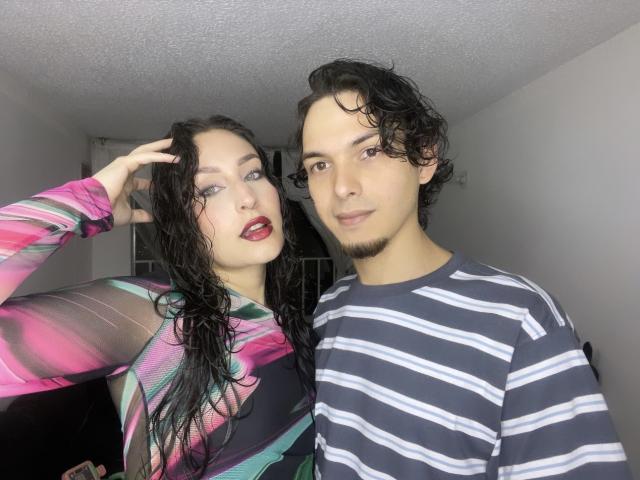 SpicyTwoDom with wet black hair, red lipstick, and colorful top, stands next to SpicyTwoDom with curly hair, striped shirt, and goatee, in a dimly lit room.