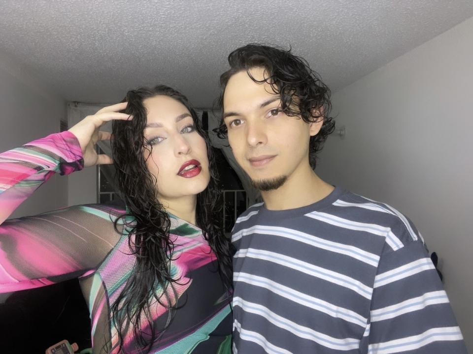 SpicyTwoDom with wet black hair, red lipstick, and colorful top, stands next to SpicyTwoDom with curly hair, striped shirt, and goatee, in a dimly lit room.