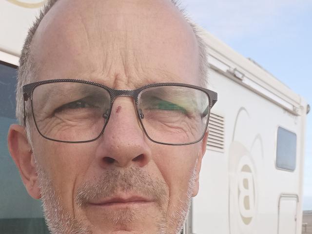 Ricky, con la testa calva, la barba grigia e i baffi, indossa occhiali neri, ha una piccola macchia rossa sul naso e un'espressione leggermente infastidita. In secondo piano, un camper bianco..