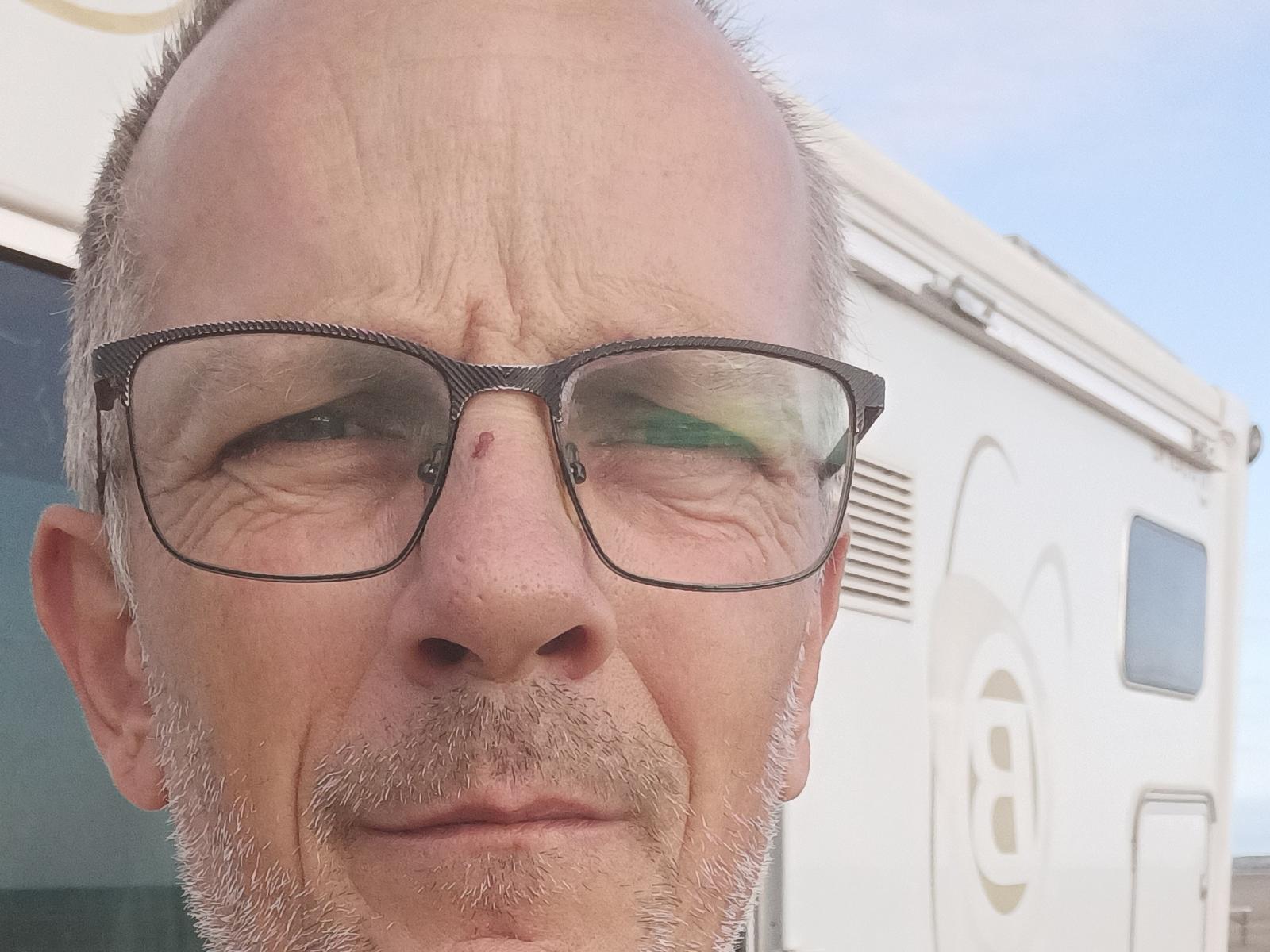 Ricky, com a cabeça raspada, barba e bigode grisalhos, usando óculos pretos, tem uma pequena mancha vermelha no nariz e uma expressão ligeiramente irritada. Uma van branca está ao fundo..