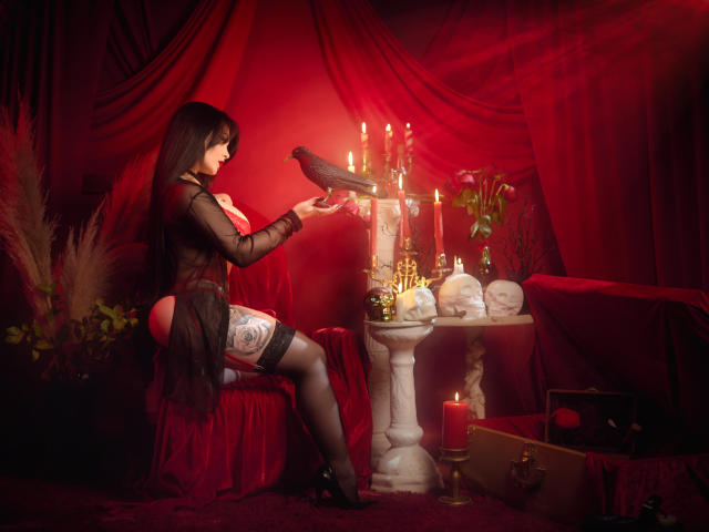 GabrielaAroms with black hair, wearing a sheer black top, exposing her tits, black stockings, and garter, holding a black bird, in a red-lit room with candles and skulls.