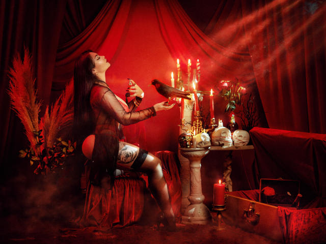 GabrielaAroms with tits and ass in sheer dress, holding bottle, feeding crow, red candles, skulls, and red curtains, sitting on red chair, surrounded by dark, moody atmosphere.