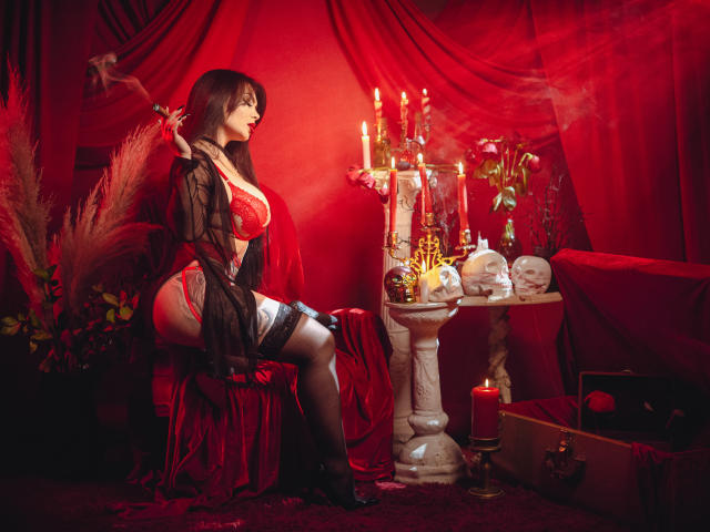 GabrielaAroms with red lace lingerie, black sheer robe, garter, and stockings, sitting on red velvet chair, holding rose, candles, skulls, and a candelabrum, red room, sexy.