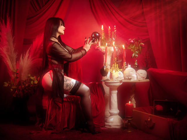 GabrielaAroms with long black hair, wearing a sheer black robe, holding a black ball, sitting on a red chair, showing her white garter, red stockings, and her ass.