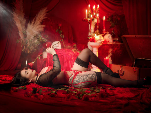 GabrielaAroms lying on red roses, wearing black sheer top, red lingerie, black stockings, holding rose stem, tattooed thigh, candles, and ornate box in dim red lighting. Boobs, ass, pussy.
