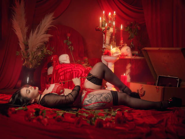 GabrielaAroms with tattoos, red lingerie, black stockings, lying on red velvet, candles, roses, and an open coffin, holding a red book, with a seductive, sexy expression.