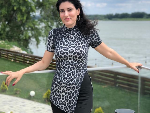 AddictedToUTS with black leopard print top, showing her tits, black pants, red nails, and hoop earrings, leaning on a glass railing, standing by a lake.