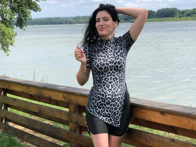 AddictedToUTS with black hair, wearing a leopard print shirt and black shorts, standing by a wooden fence, hand in hair, smiling, showing her tits, by a lake.
