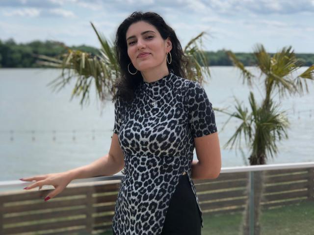 AddictedToUTS with medium tits, wearing a leopard print dress, standing by a wooden fence, palms in background, hoop earrings, red nails, and a slight smile, under a cloudy sky.