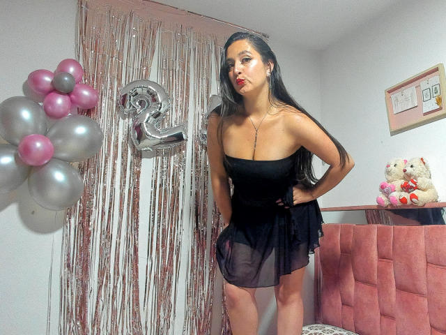 VenusSweetPalmer, con cabello negro largo, vestida con un vestido negro transparente, labios rojos y sensuales, con las manos en las caderas, de pie en una habitación con globos rosas, el número 2 en color plateado y un oso de peluche..