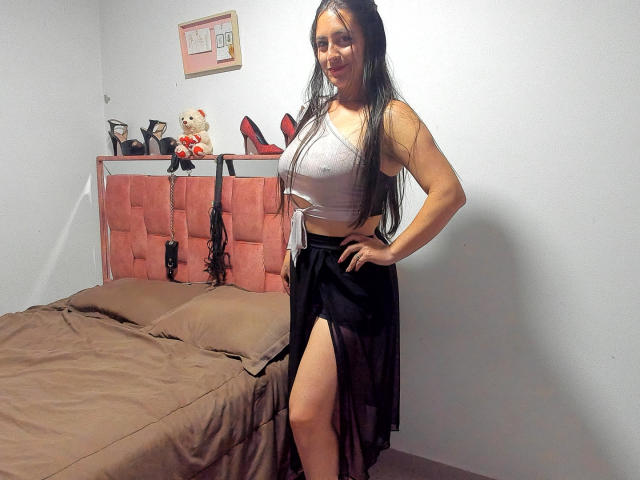 VenusSweetPalmer with long black hair, white crop top, and black skirt with thigh-high slit, standing in bedroom, showing off her big ass and small tits.