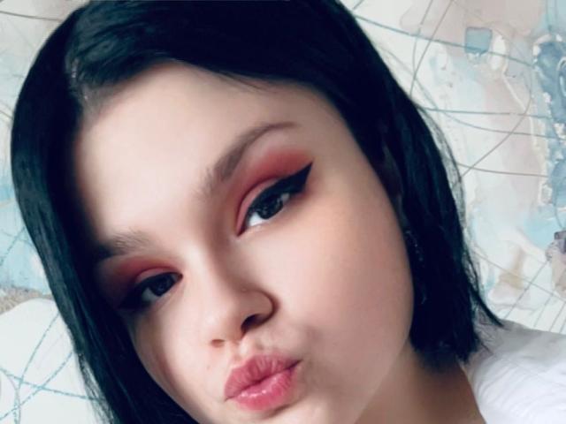 AdelinaOlsen has pale skin, black straight hair, and pinkish-red eyeshadow with bold black eyeliner. She's pouting her pink lips, looking slightly to the side.