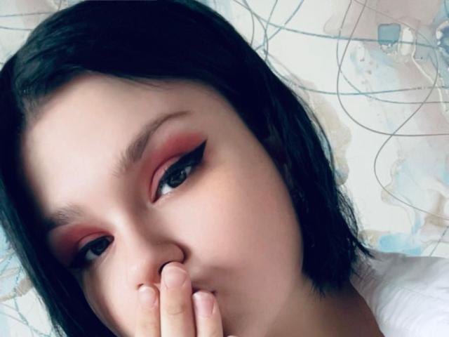 AdelinaOlsen's face with black shoulder-length hair, pink and red eyeshadow, black eyeliner, and her finger covering her nose. She has fair skin and dark eyes.