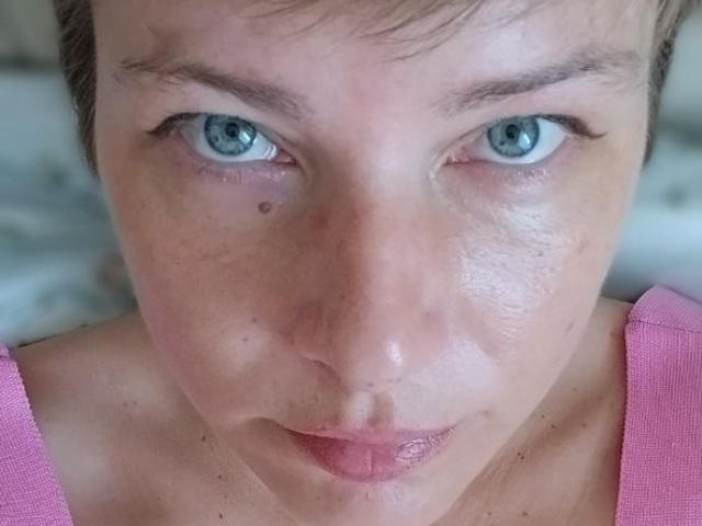 MissYesminet has blue eyes, a small mole on her right cheek, and is wearing a pink top. Her face is slightly shiny with subtle freckles and short brown hair.