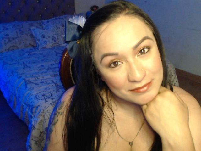 LucianaDia with long black hair, brown eyes, and a slight smile, resting her chin on her hand, showing a tattoo on her right arm, and wearing a necklace.