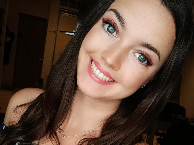 Vanilia69 with blue eyes, dark brown hair, pink lipstick, and red eyeshadow, smiling at the camera, showing white teeth, wearing a necklace, in a brightly lit room.