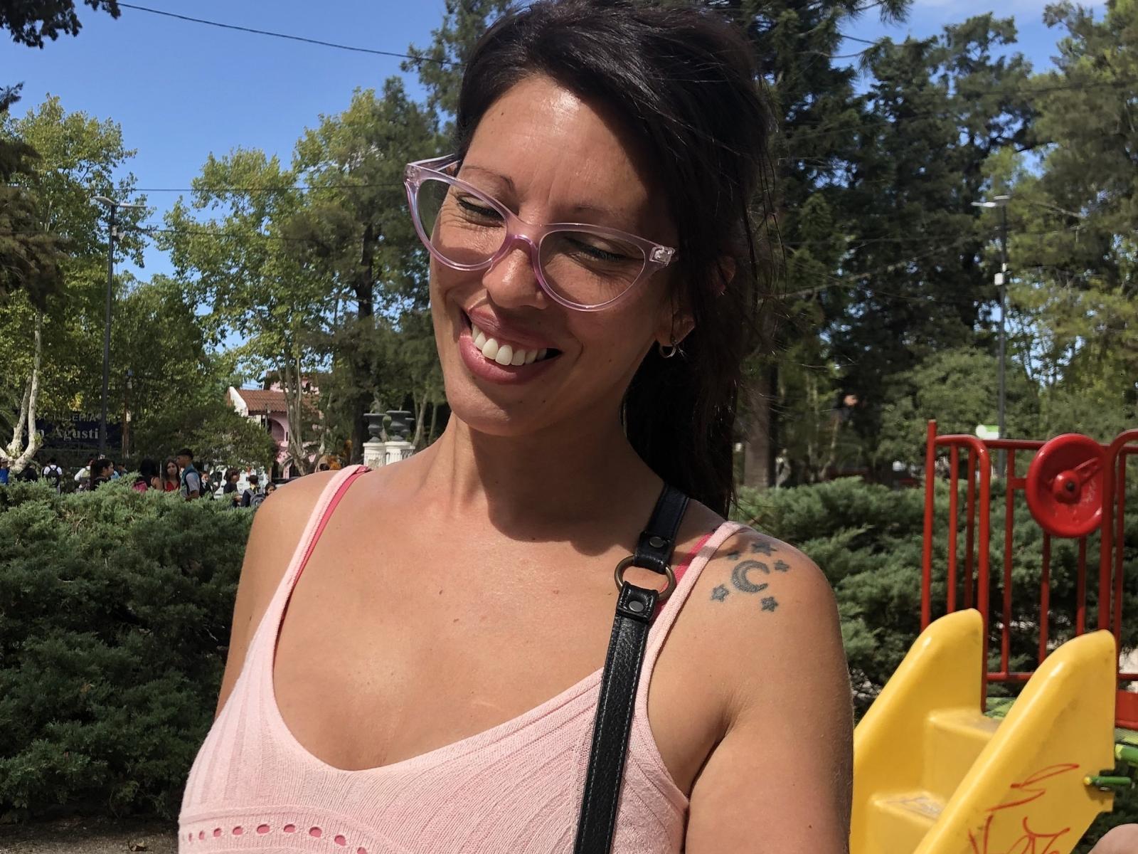 MargotMilf with pink glasses, smiling, pink tank top, black strap, tattoo on right shoulder, sunny day, trees, yellow playground structure, brown hair, medium tits, cute, happy.