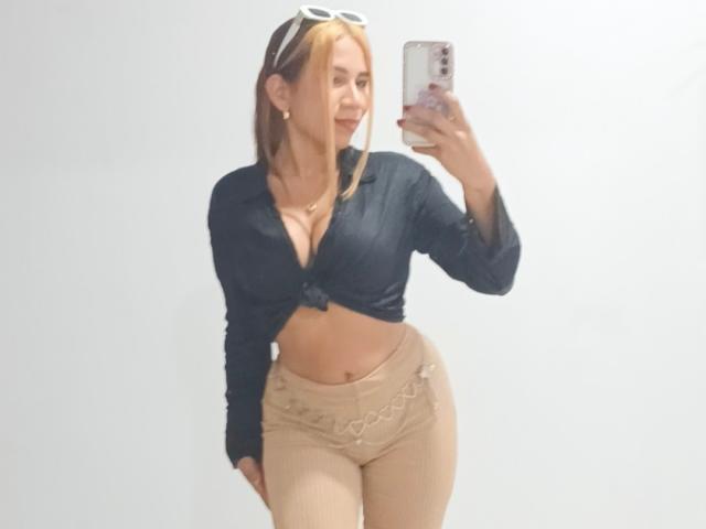 LunaMiranda, avec ses cheveux blonds, portant une chemise noire nouée qui met en valeur sa poitrine généreuse, un pantalon beige taille haute et des lunettes de soleil sur la tête, prend un selfie avec un smartphone..