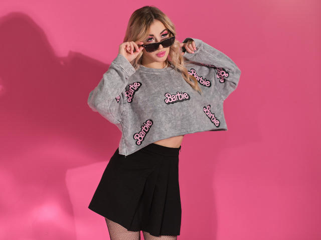HelenElleson with blonde hair, wearing a gray "Barbie" crop sweater, black skirt, fishnet stockings, and black sunglasses, standing against a pink background, holding her sunglasses.
