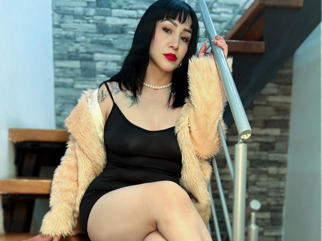 MartinaMilf com cabelo preto, batom vermelho, vestido preto, colar de pérolas e casaco de pele, sentada nas escadas, segurando o corrimão, mostrando tatuagens e exibindo sua bunda..