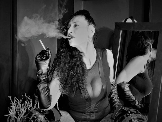 KarineDuponts with curly hair, smoking a cigarette, wearing a low-cut top that shows her big tits, black gloves, and a mirror reflecting her back. Her ass is visible.
