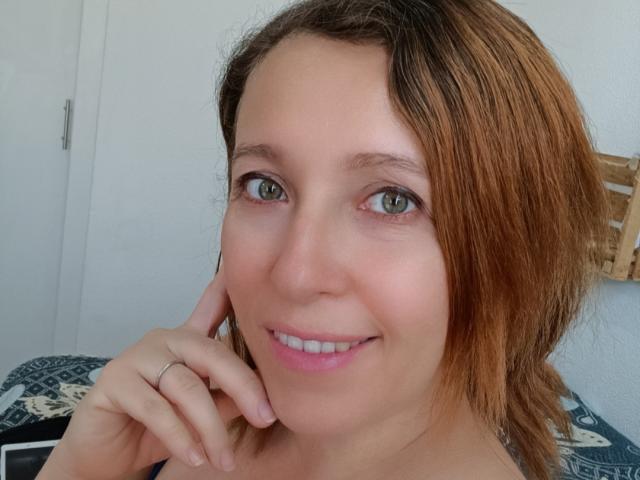 CharlottePonze has light green eyes, a slight smile, and is touching her chin with her right hand. She has light brown, slightly wavy hair and is wearing a ring.