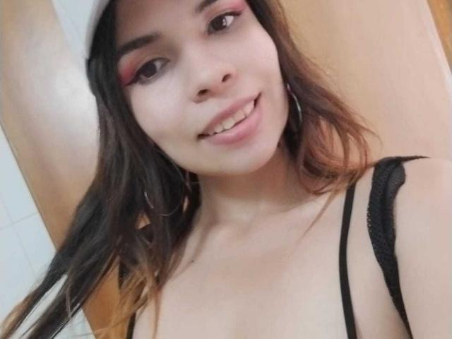 VioletaStone with long brown hair, pink eyeshadow, black straps, smiling, small tits, hoop earrings, light skin, arm raised, wearing a black top, taking a selfie.