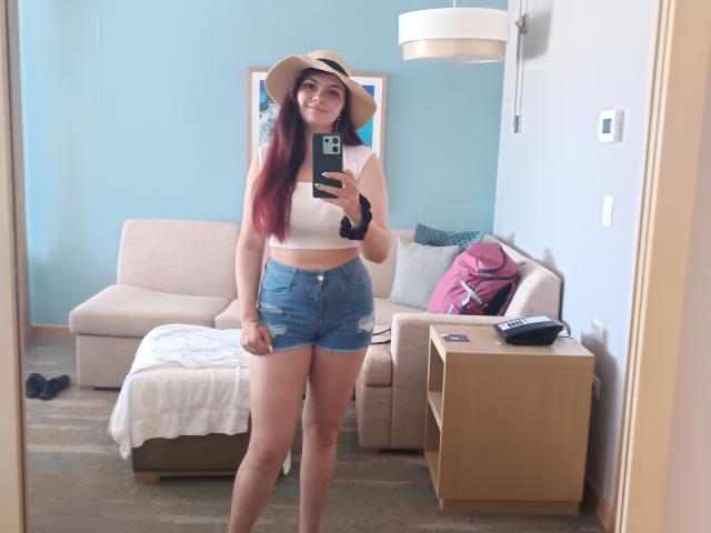 KatherinLatin with red-tinted long hair, wearing a white crop top, denim shorts, and a sun hat, taking a mirror selfie in a room, showing her ass and tits.
