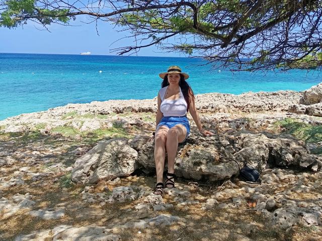 Katherin, in versione latina, con il seno scoperto, indossa una canotta bianca, pantaloncini di jeans e un cappello da sole, seduta su delle rocce vicino al mare blu, con sandali neri, sotto un albero..
