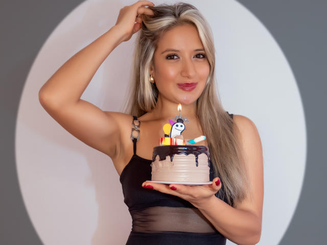 ValeriaHunter69 with blonde hair, black dress, holding a chocolate cake with candles, smiling, right arm raised, red nails, white background, cute, sexy, cake on her left hand.