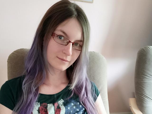 EmmaLust with purple ombre hair, glasses, green shirt, small tits, fair skin, and a slight smile, sitting in a chair.