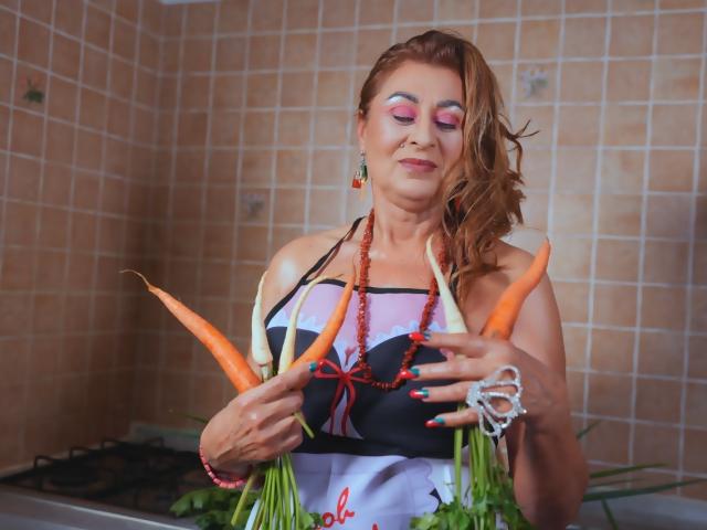 KarlaHartley, middle-aged woman with red hair, holding large carrots in kitchen, wearing black and pink top, red bead necklace, pink eye makeup, smiling, visible tits, green nails.