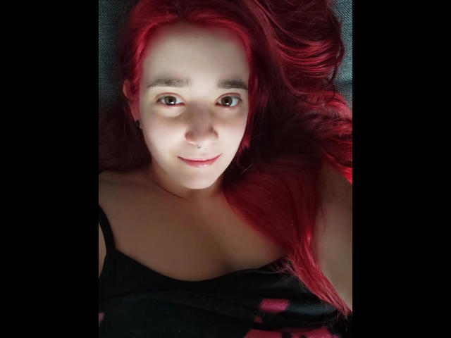 LadyJosefine-hot with red hair, fair skin, brown eyes, nose ring, and black top, lying down, smiling softly. Boobs visible, black top with pink pattern, head slightly tilted.