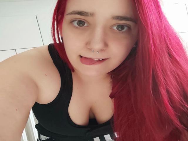 LadyJosefine-hot with red hair, nose piercing, and tongue out, wearing a black tank top, showing her boobs, pale skin, and green eyes, in a selfie.