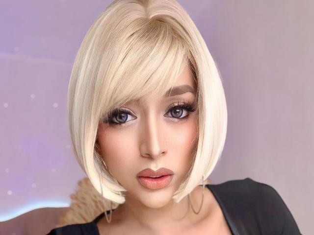 YourDreamGoddess with blonde bob, blue eyes, heavy eyeliner, and pink lips, wearing black top, looking straight at camera, has cute face and soft makeup.