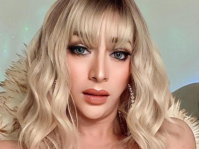 Blond YourDreamGoddess with wavy hair, blue eyes, and full lips, wearing fur, with subtle makeup and diamond earrings, stares intensely at the camera.