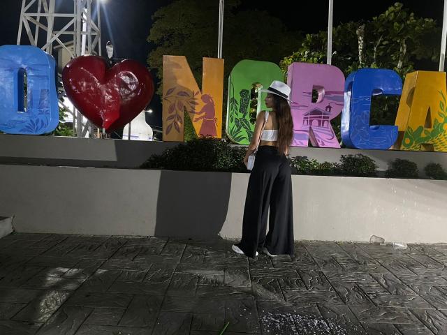 ElenaSantini with long brown hair, white hat, white strap top, black high-waisted pants, standing in front of colorful, large letters, on a tiled, dark sidewalk at night.