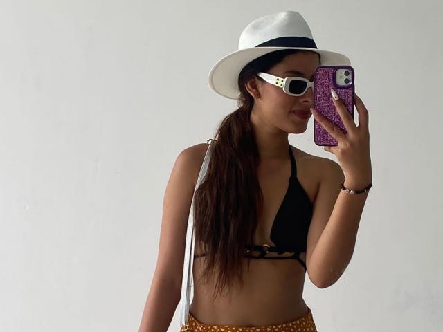 ElenaSantini, wearing a white hat, black bikini top, and orange polka-dot bottom, takes a mirror selfie with a purple glitter phone, showing her tits and long brown ponytail.