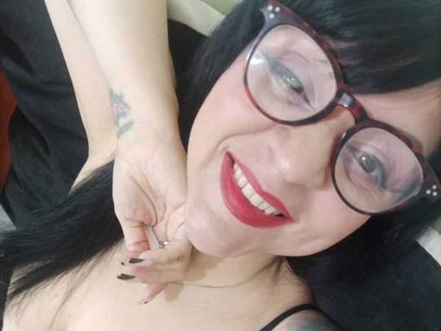 AlishX with black hair, red lipstick, and glasses, smiling, showing a tattoo on her hand, touching her neck, wearing a black top, with black fingernails.