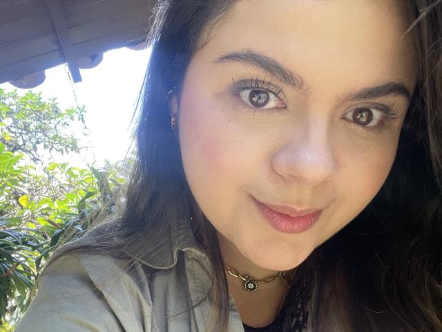 MissSofiia's face with brown eyes, long eyelashes, and a slight smile, wearing a gray shirt, black necklace, and pink lipstick, under a shaded area with green foliage.