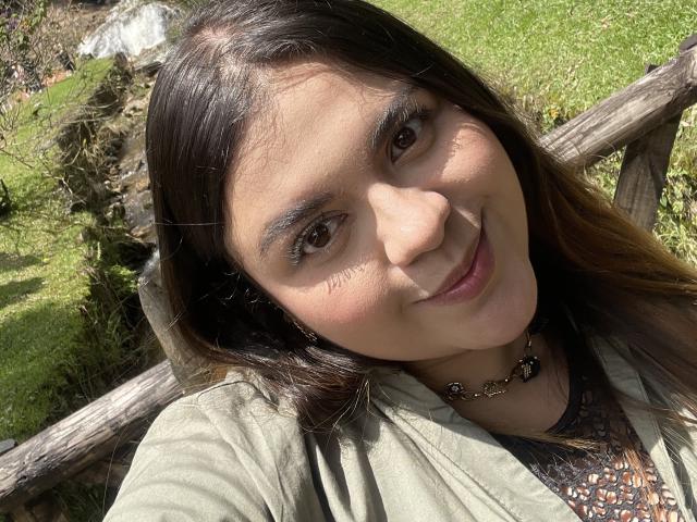 MissSofiia's face with brown eyes, long dark brown hair, green shirt, and necklace, smiling in sunlight, showing her cute nose and subtle makeup. Her ass is not visible.