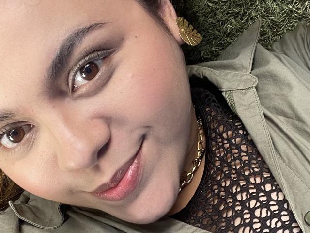 MissSofiia's face, showing brown eyes, subtle makeup, and a slight smile. She's wearing a green jacket, black mesh top, gold chain, and green leaf earrings. Her pussy isn't visible.