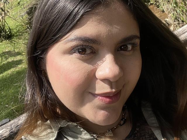 MissSofiia's face, showing brown eyes, straight brown hair, light skin, and a subtle smile, with a small nose and pink lips, wearing a greenish patterned shirt, sunlight on her face.