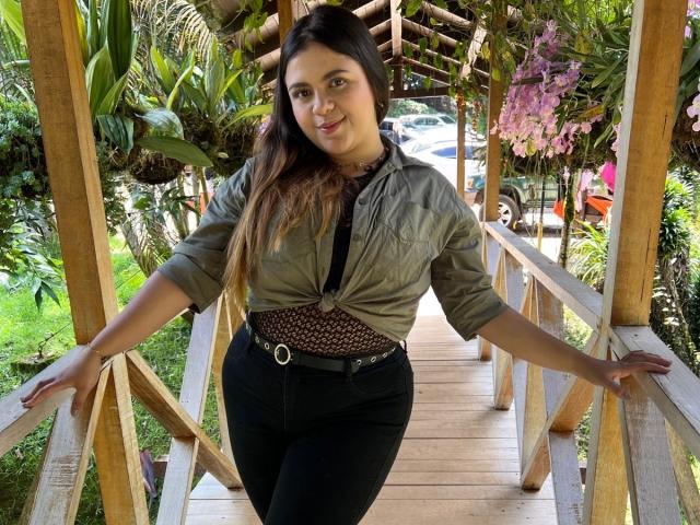 MissSofiia stands on a wooden bridge, arms out, wearing a tied green shirt, black pants, and a patterned top, with long brown hair and a cute smile.