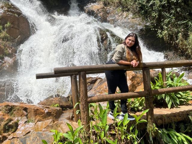 MissSofiia leans on a wooden railing, smiling, with a waterfall behind her. She's wearing a green jacket, black pants, and white shoes, with long brown hair.