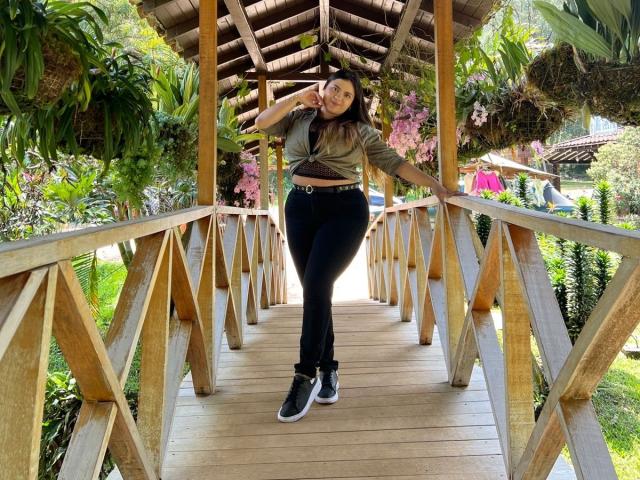 MissSofiia with big ass, tied-up green top, black pants, and black sneakers, standing on wooden bridge, hand in hair, hanging plants, and wooden roof overhead.